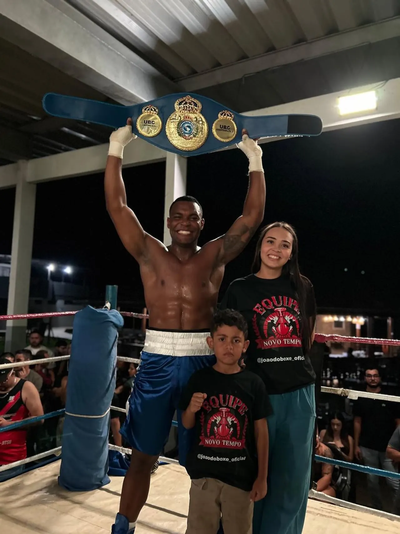 João do Boxe no ringue, erguendo o cinturão UBC, ao lado da esposa e de um menino com a camisa da Equipe Novo Tempo